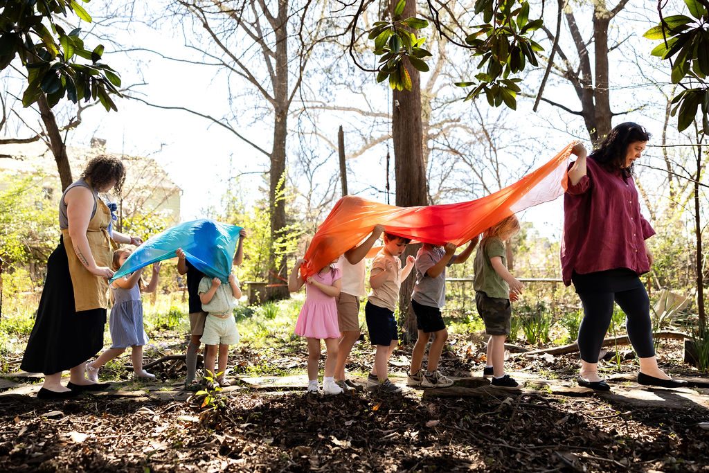 Sally and a teacher lead children under flowing silks through a garden